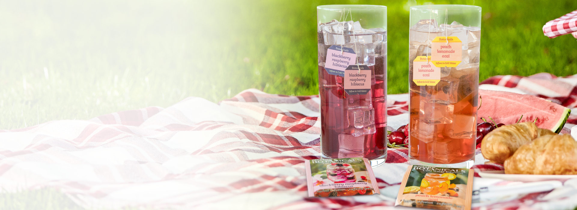Two glass tumblers with fruit infusions on a picnic blanket with snacks and a watermelon.