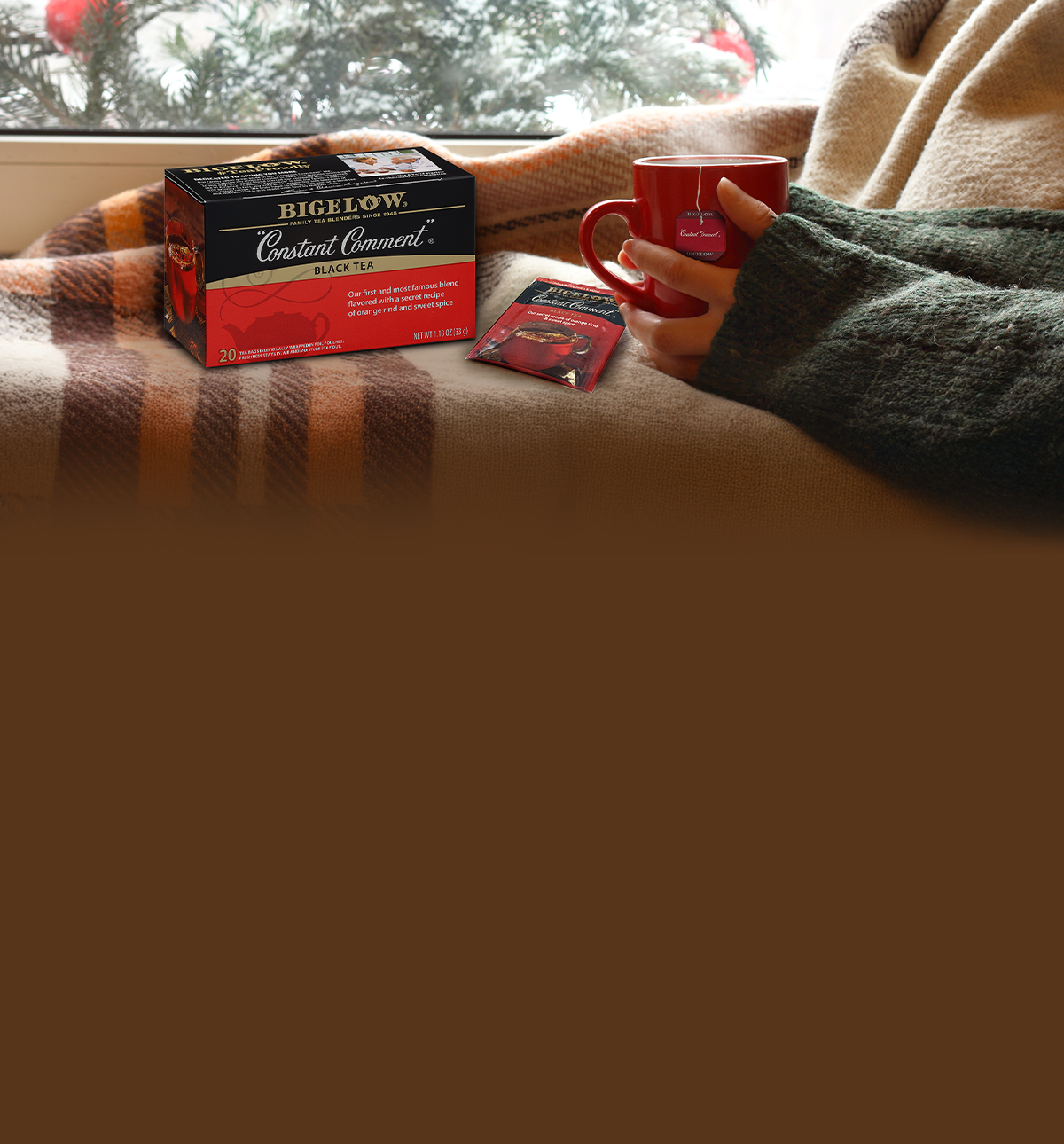 Person holding a red mug with Bigelow tea packaging on a couch
