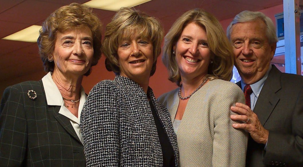 Four people posing together indoors. Two women and one man on the left wear dark blazers, while a woman on the right wears a light beige cardigan.