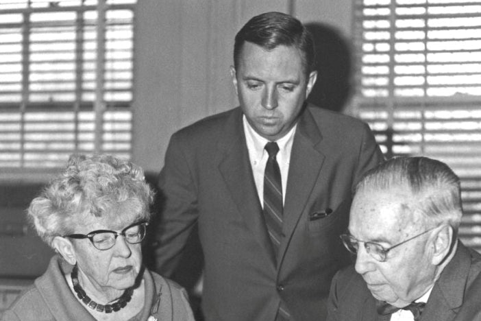 Black and white photograph of three people in a meeting setting, with an older woman on the left wearing glasses and a cardigan, a man standing in the center in a suit and tie, and an older man on the right in a suit and bow tie looking at papers at a desk.