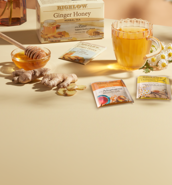 A styled product shot of Bichow Ginger Honey Herbal Tea with its box, a glass mug of amber tea, honey, ginger root, chamomile flowers, and three single-serve packet packs on a warm beige surface.
