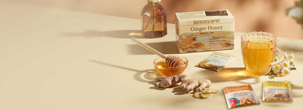 A boxed tea product labeled Bigelow Ginger Honey sits on a warm beige tabletop with a glass of amber tea, ginger root pieces, honey in a small bowl, and several tea sachets arranged around it. A blurred human figure is visible in the background, and the scene is styled with natural light and a soft neutral backdrop.