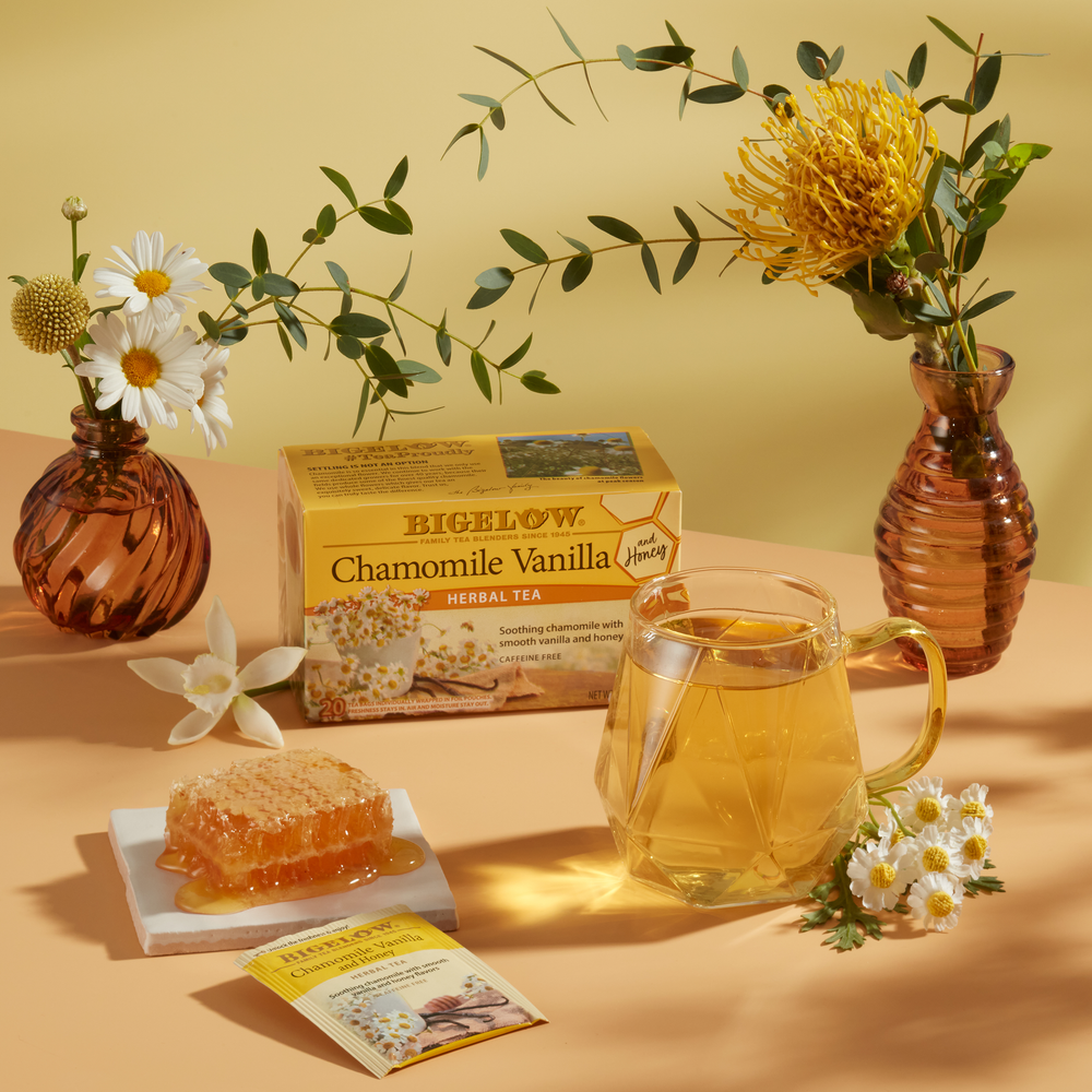 Glass cup of Chamomile Vanilla and Honey Herbal Tea with box and foil packet on table with flowers in 2 vases