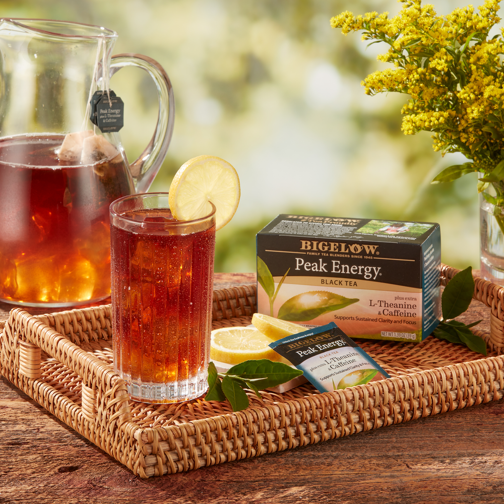 Glass pitcher and glass of  Peak Energy Plus Extra L-Theanine and Caffeine Black Tea over ice with box and foil packet on tray with flowers in vase
