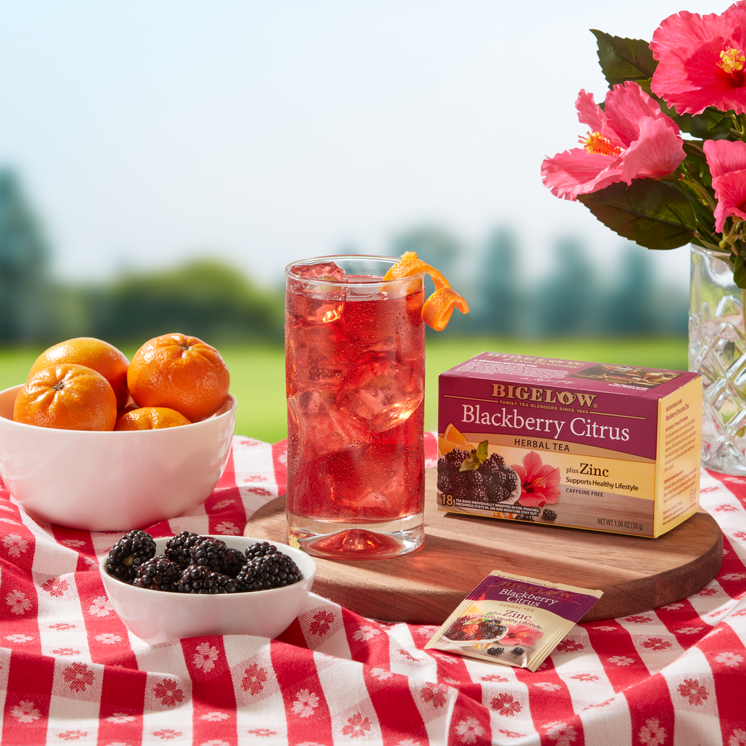 Glass of Iced Blackberry Citrus Herbal Tea Plus Zinc with box of tea in a picnic setting with a bowl of fruit