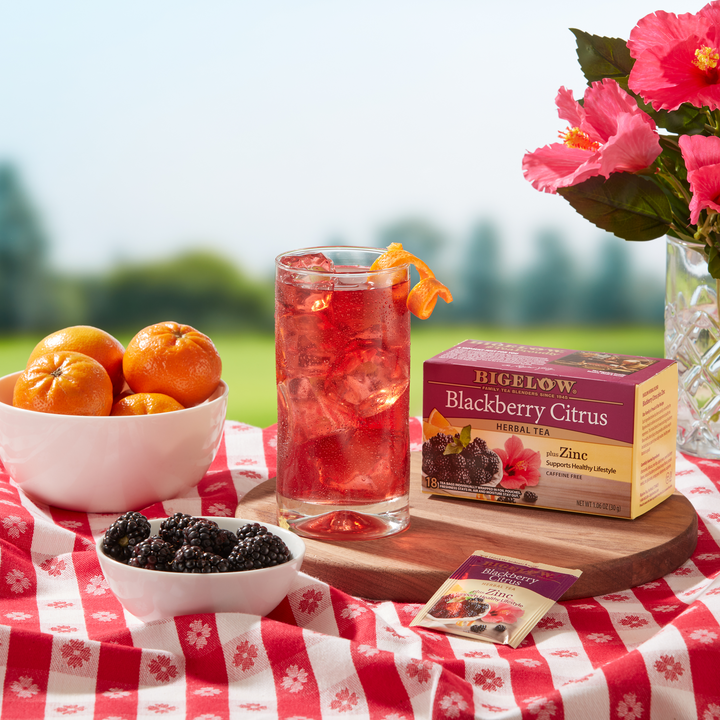Glass of Iced Blackberry Citrus Herbal Tea Plus Zinc with box of tea in a picnic setting with a bowl of fruit