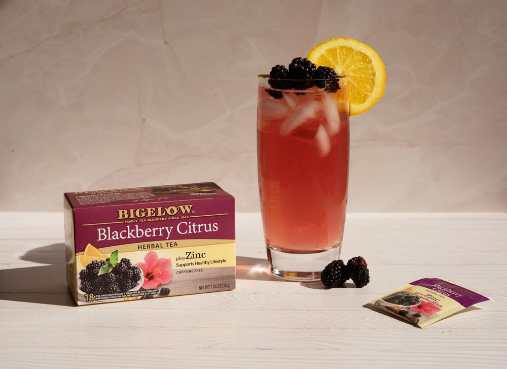 Bigelow Blackberry Citrus herbal tea box with a glass of iced tea, blackberries, and an orange slice on a light surface.