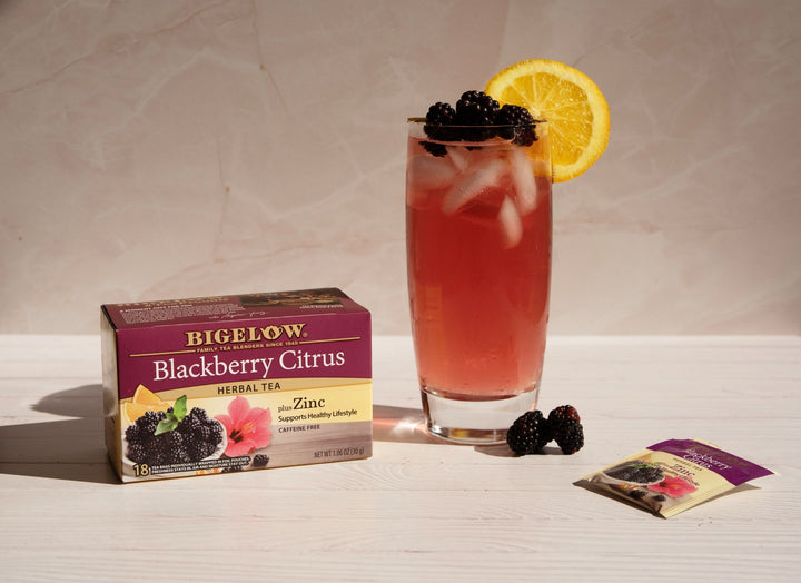 Bigelow Blackberry Citrus herbal tea box with a glass of iced tea, blackberries, and an orange slice on a light surface.
