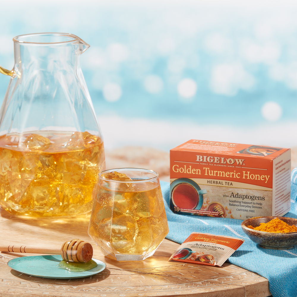 Glass and pitcher of Bigelow Golden Turmeric Honey Herbal Iced Tea with turmeric in bowl with honey dripper with a seaside setting
