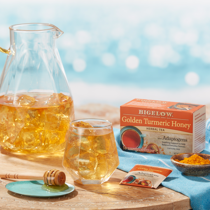 Glass and pitcher of Bigelow Golden Turmeric Honey Herbal Iced Tea with turmeric in bowl with honey dripper with a seaside setting
