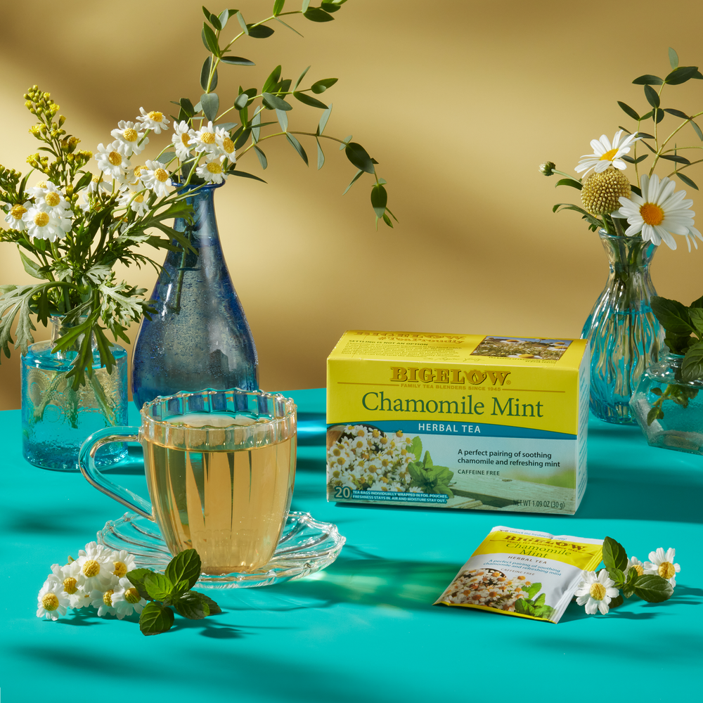 Box of Chamomile Mint Herbal Tea  with glass mug and foil packets on teal tablecloth with flowers in vase